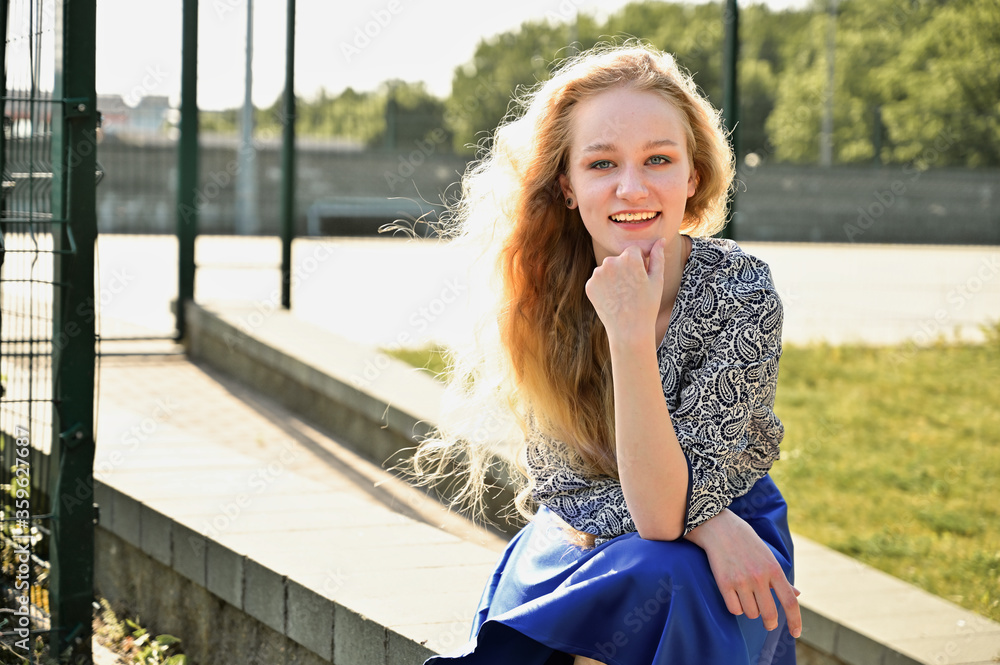 Photo of a caucasian blonde in a park in sunny weather. Girl sitting on a stand and smiling happy