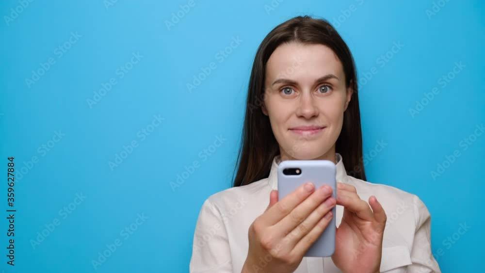 Portrait of beautiful calm brunette girl stands with mobile phone, wears white shirt, points aside with index finger on blank space, isolated on blue studio background. People, religion, technology Portrait of beautiful calm brunette girl stands with mobile phone, wears white shirt, points aside with index finger on blank space, isolated on blue studio background. People, religion, technology