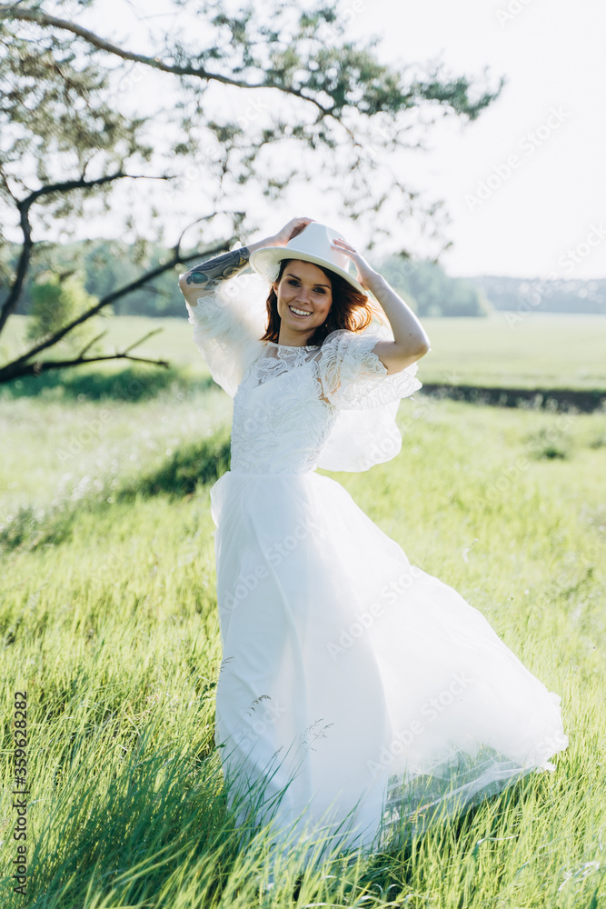 stylish woman in fedora hat  white dress walking in the field with trees