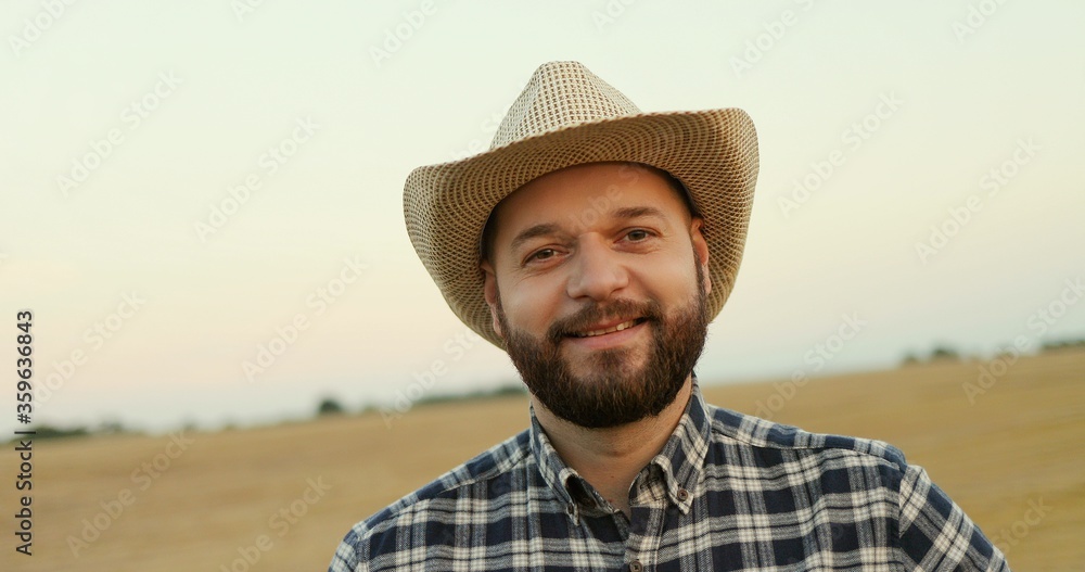 Portrait shot of the young farmer in a hat looking at the side and then smiling to the camera in the field. Close up.