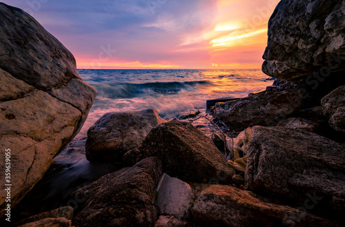 Ocean water splash on rock beach with pink and golden sunset sky. Sea wave splashing on stone at sea shore on summer. Tires and plastic sacks on tropical beach. Beach and ocean pollution. Soft wave.