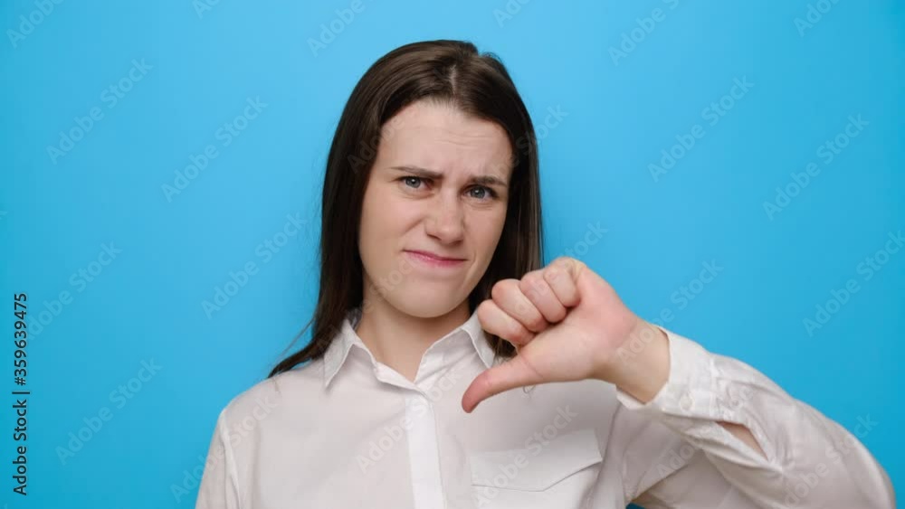 Unhappy tired sad young girl in white shirt, isolated on blue background. Upset woman 20s 30s years old looking disapprovingly at camera showing thumb down. People sincere emotions lifestyle concept Unhappy tired sad young girl in white shirt, isolated on blue background. Upset woman 20s 30s years old looking disapprovingly at camera showing thumb down. People sincere emotions lifestyle concept