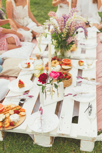 Summer celebration picnic for friends in the garden. Tasty food and cold drinks. Beautiful flowers decoration. Pallet table. Hen-party