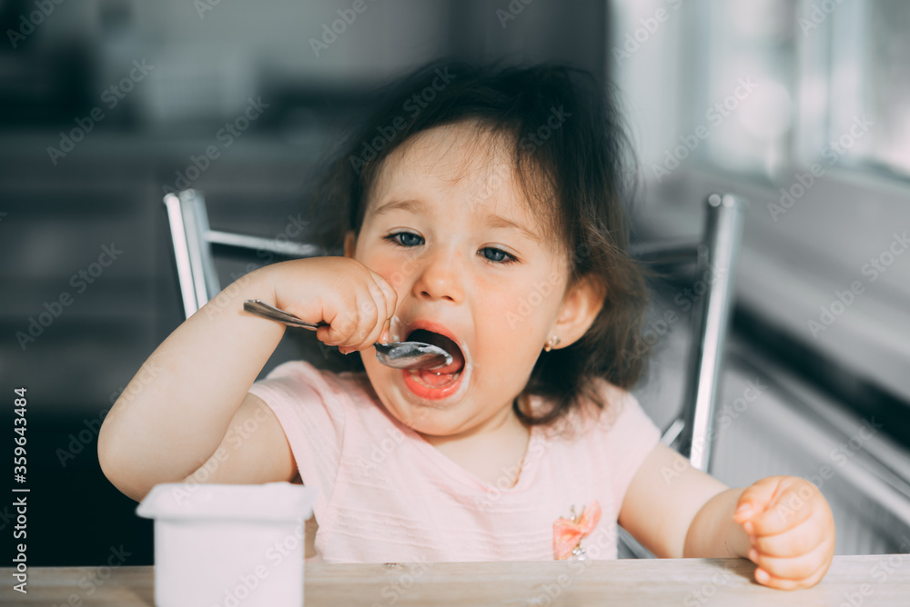 Cute, funny baby girl eating yogurt in the kitchen in a pink dress in the afternoon Cute, funny baby girl eating yogurt in the kitchen in a pink dress in the afternoon