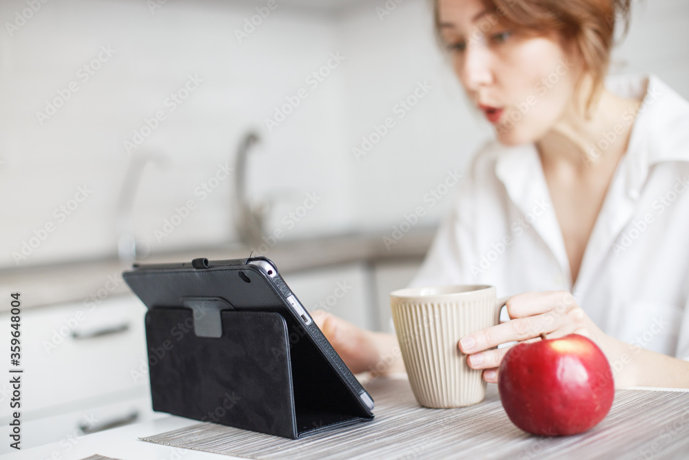 Fototapeta premium Happy surprised woman with cup of coffee or tea using laptop in the kitchen in white shirt. girl with a tablet