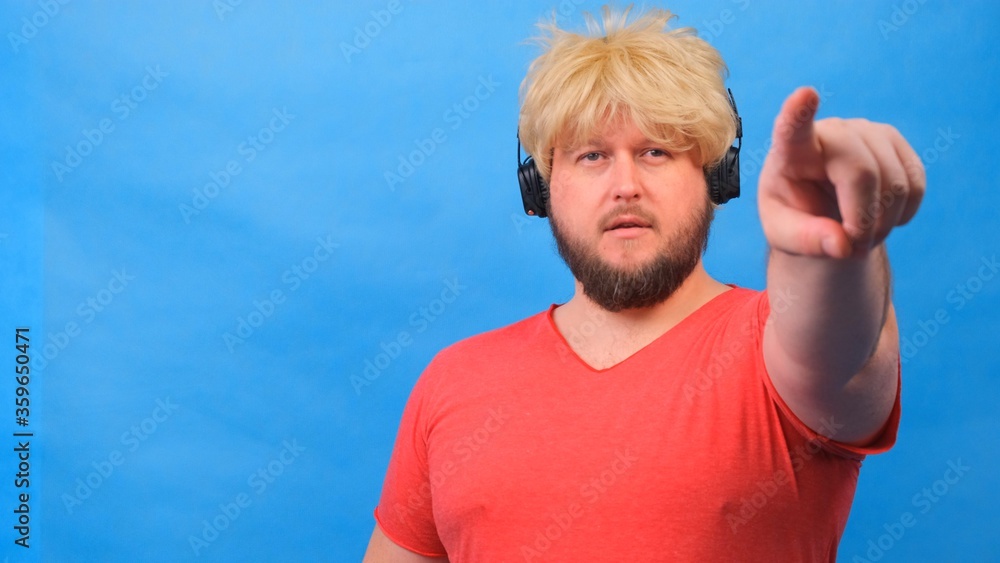 Naklejka premium freaky fat man in a wig and a pink T-shirt in headphones shows a finger on the camera at you against a blue background.