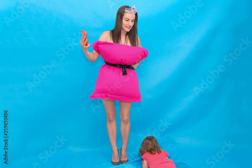 woman in pink pillow dress and crown take a selfie photo on a mobile phone for a crazy Pillow Challenge on a blue background due to being at home isolated, a child is crying nearby. Fashionable girl photo