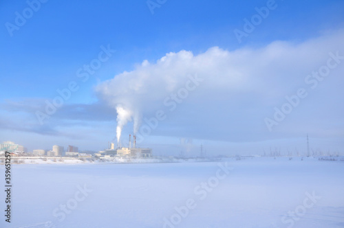 Thermal power station with the steam from the pipes, producing heat energy, view over blue morning foggy sky during cold winter with frozen river on the foreground