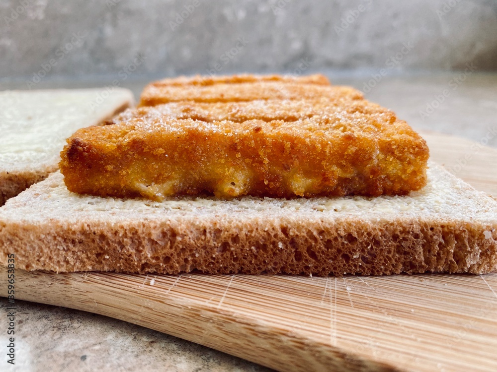 Closeup view of crispy fish fingers covered in salt and vinegar on buttered bread in the kitchen. Breaded fish finger sandwich preparation on wooden board.