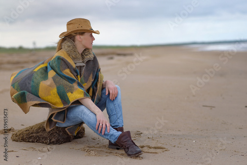 Murais de parede woman sitting on the sand by the sea, dressed in a poncho and in a koyboy hat