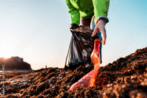 A woman in a green jacket, with a garbage bag in her hands, reaches for a bottle to the ground. Bottom view. The sky is in the background. Close up and copy space. Concept of environmental pollution