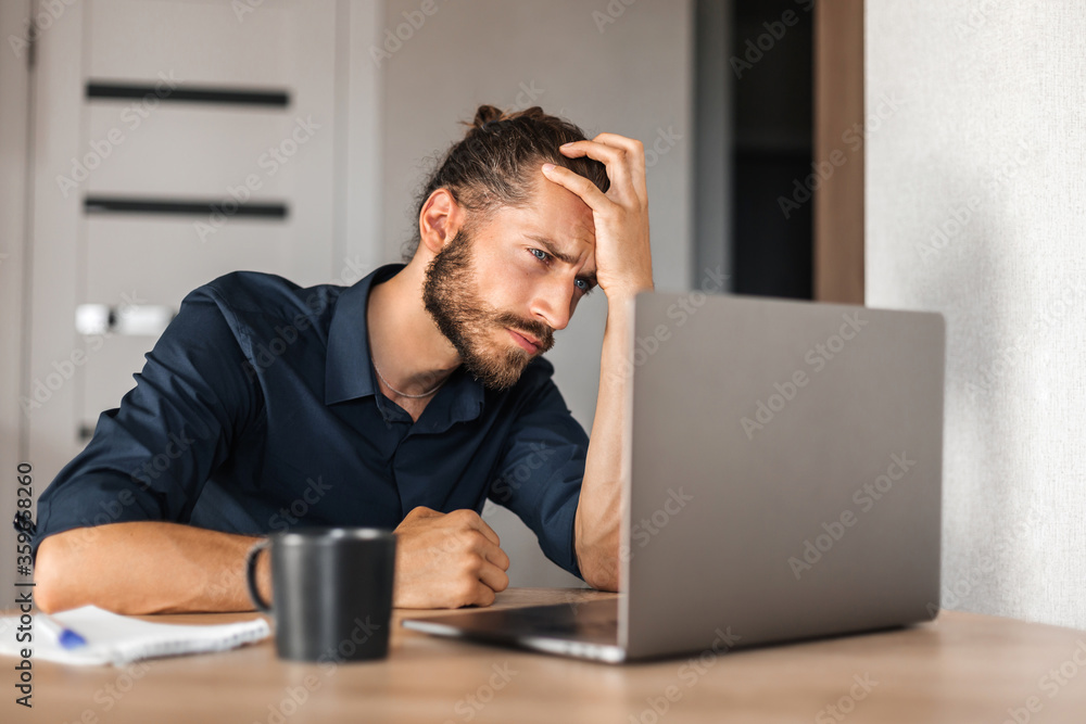A tired man is looking at a laptop. Young man holding his head. Work at home. Young man tired of work A tired man is looking at a laptop. Young man holding his head. Work at home. Young man tired of work