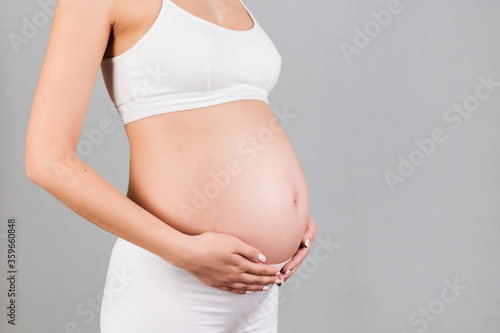 Cropped image of pregnant woman in white underwear touching her belly at gray background. Future mother is waiting her baby. Copy space