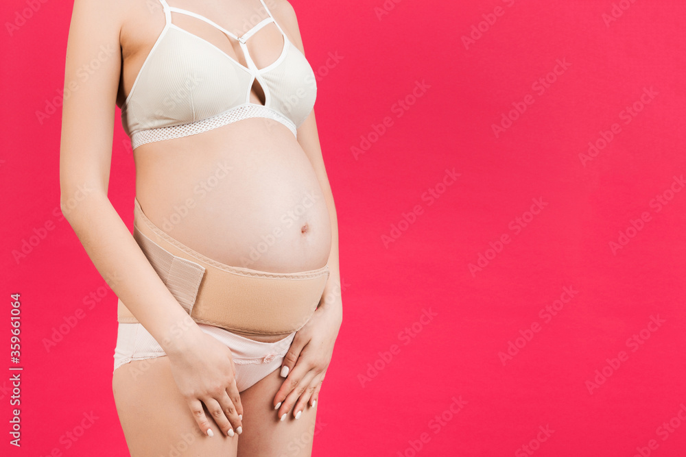 Close up of supporting orthopedic bandage against backache on pregnant woman in underwear at pink background with copy space. Orthopedic abdominal support belt concept Close up of supporting orthopedic bandage against backache on pregnant woman in underwear at pink background with copy space. Orthopedic abdominal support belt concept