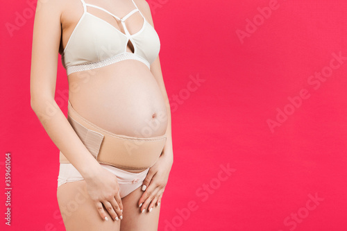 Close up of supporting orthopedic bandage against backache on pregnant woman in underwear at pink background with copy space. Orthopedic abdominal support belt concept