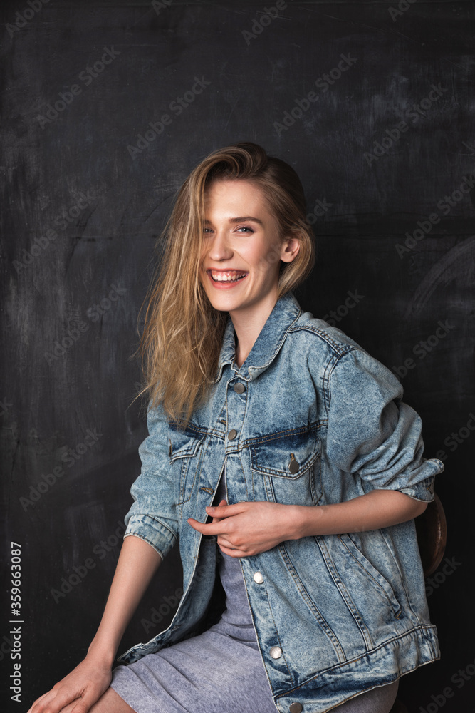 Blonde woman in a blue jeans jacket and grey dress smiling and looking at the camera. A black chalkboard on the background Blonde woman in a blue jeans jacket and grey dress smiling and looking at the camera. A black chalkboard on the background