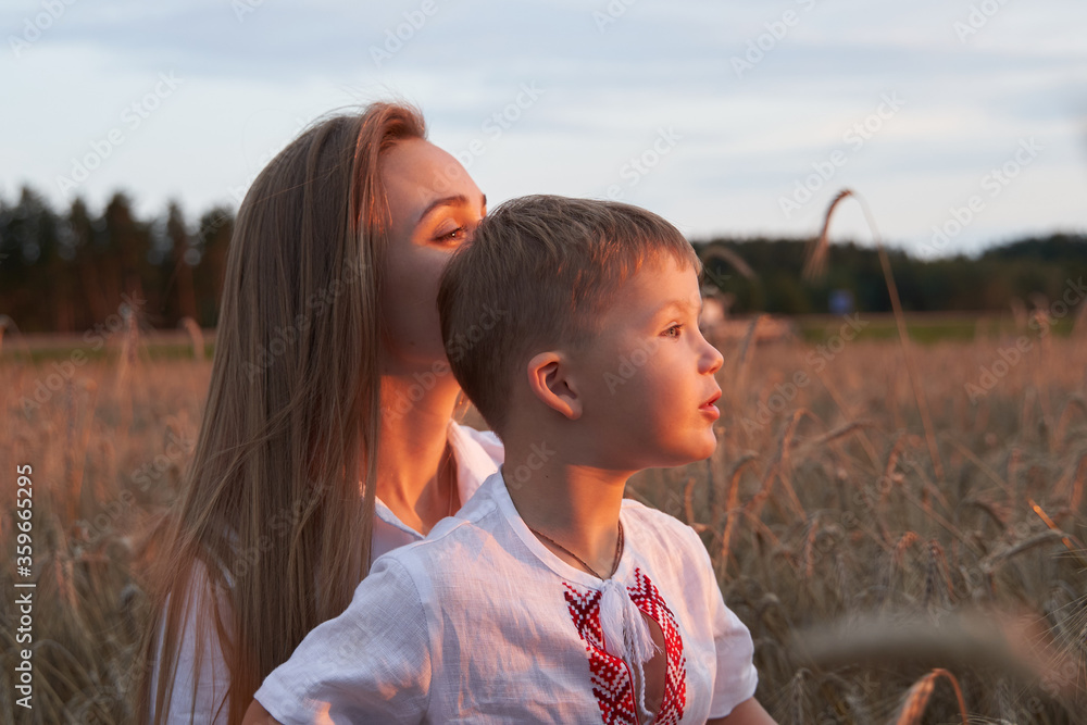 Fototapeta premium Portrait of cheerful son with camera sitting on mothers laps amidst wheat field