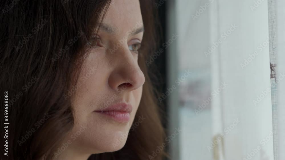 Portrait of beautiful woman standing by the window at home, attractive female face closeup, natural human beauty, brunette woman with long hair looking out the window. Portrait of beautiful woman standing by the window at home, attractive female face closeup, natural human beauty, brunette woman with long hair looking out the window.