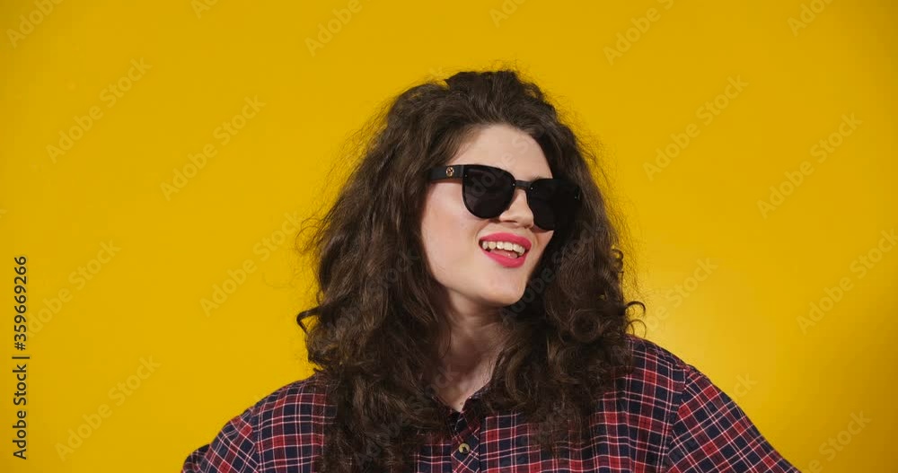Close-up attractive plus-size model posing. Curly-haired brunette on a bright yellow studio background demonstrating different situations. Trendy smiling attractive girl wearing dark sunglasses Close-up attractive plus-size model posing. Curly-haired brunette on a bright yellow studio background demonstrating different situations. Trendy smiling attractive girl wearing dark sunglasses