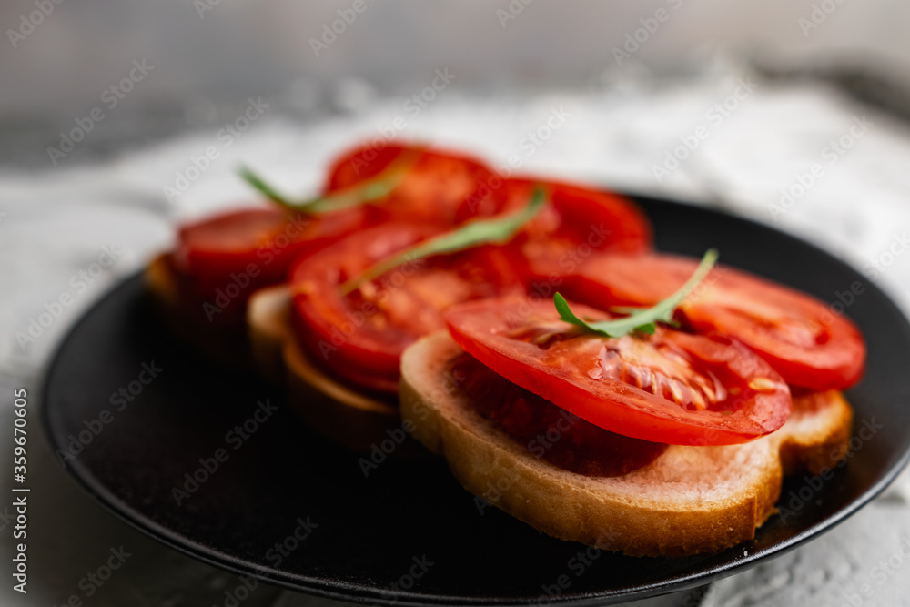Three sandwiches with fresh and bright red tomatoes and smoked sausage, decorated with a leaf of green arugula on a matte black plate and a white stone background Three sandwiches with fresh and bright red tomatoes and smoked sausage, decorated with a leaf of green arugula on a matte black plate and a white stone background