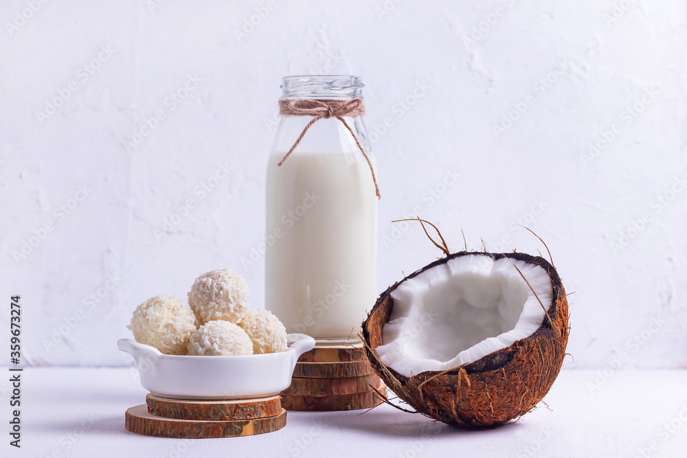 Bottle of coconut vegan milk on wooden coasters with chopped brown coconut and round sweets with coconut flakes on a white background Bottle of coconut vegan milk on wooden coasters with chopped brown coconut and round sweets with coconut flakes on a white background