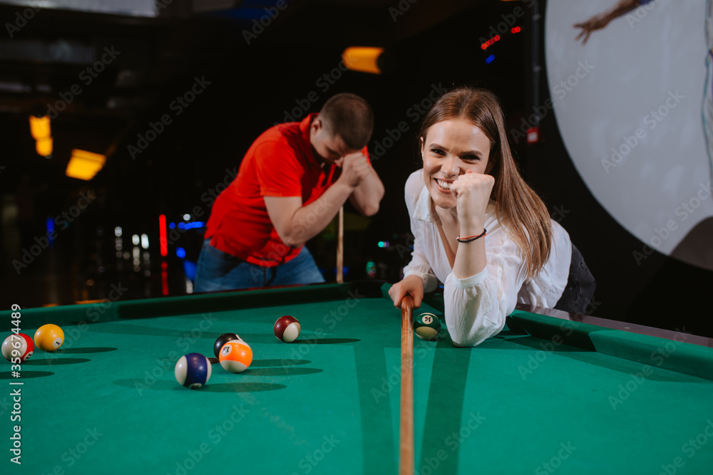 Fototapeta premium A young caucasian woman and a caucasian man play billiards. A beautiful woman is aiming a ball, while a man in a red T-shirt is standing next to her. Fun time
