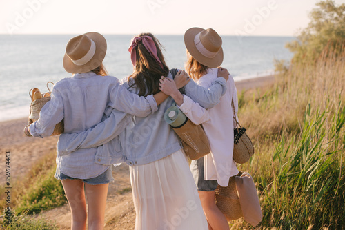 Three young beautiful women female friends going to have summer picnic on a beach at sunset.