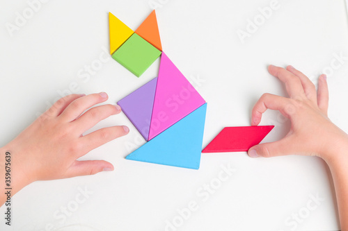 Tangram colored geometric puzzle pieces with childs hands moving pieces on white table, top view. 