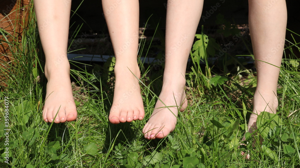two pairs of bare feet of children sitting on a bench and touching fresh green grass with their feet on a warm sunny summer day