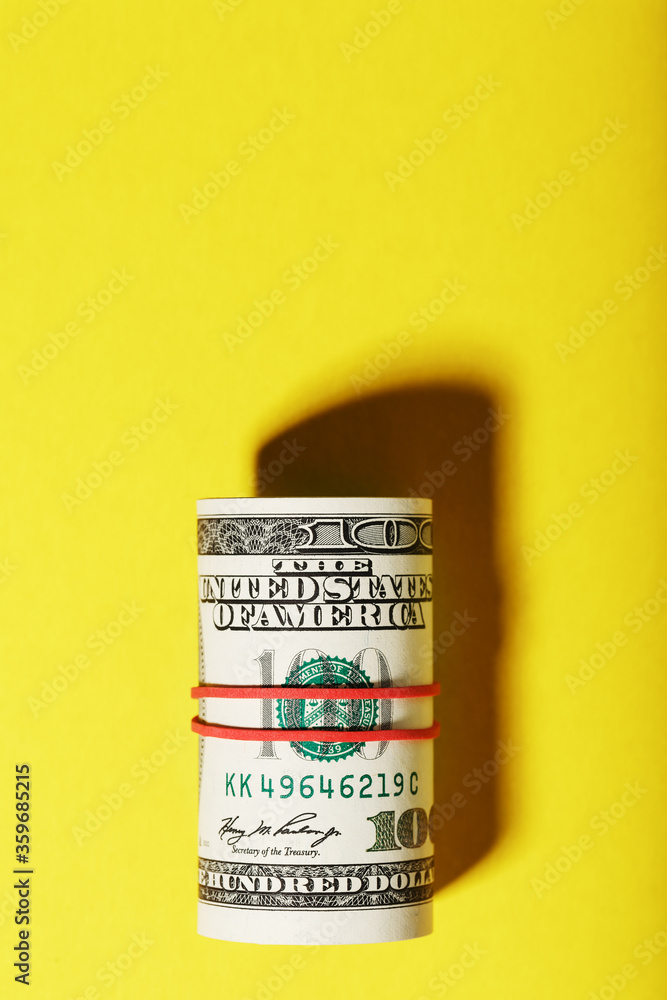 A roll of hundred-dollar American bills is tied with a red elastic band on a yellow background. A roll of hundred-dollar American bills is tied with a red elastic band on a yellow background.
