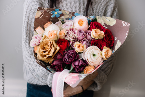 Very nice young woman holding beautiful blossoming bouquet of roses  ranunculus  carnations  tulips flowers cream pink and brown in colors on the grey background