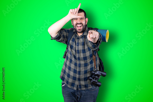 Obraz na plátne Closeup portrait serious young traveler bearded man showing loser sign on forehead looking at you with disgust at camera isolated green studio background