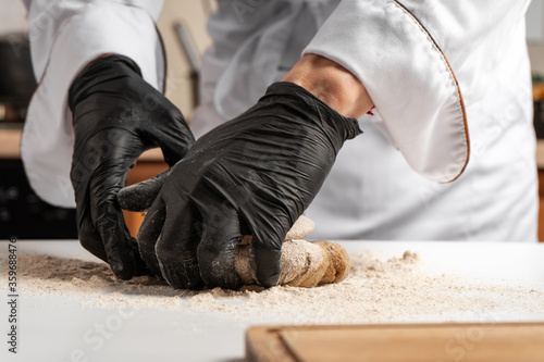 The chef in black gloves kneads dough from wheat flour on a white table