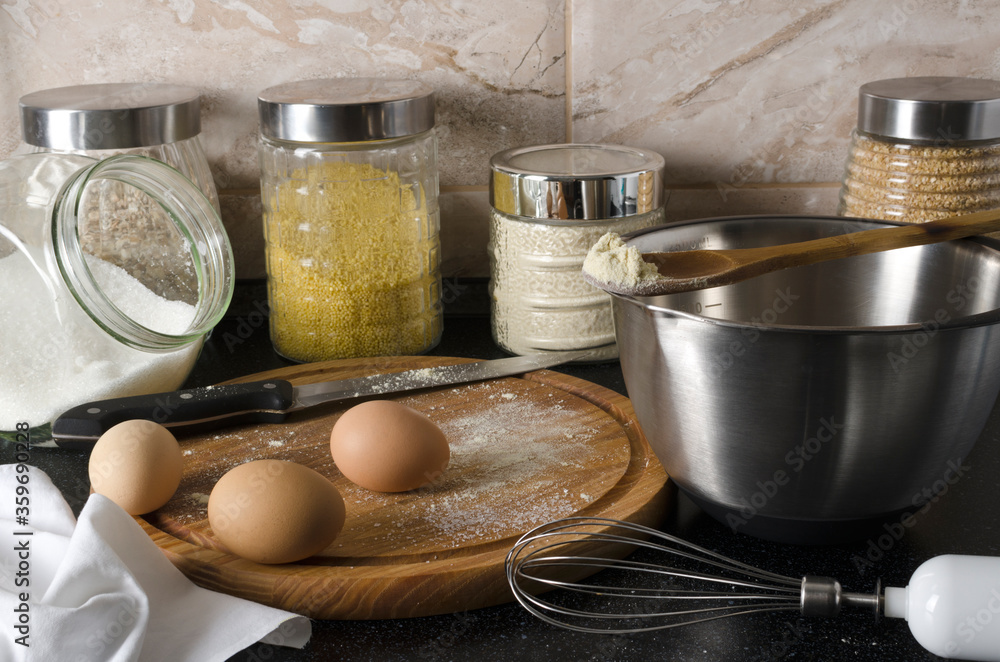 Process of cooking. Closeup of wooden board, eggs, flour, glass jars with cereals, steel bowl, sugar and whisk on the black table against wall Process of cooking. Closeup of wooden board, eggs, flour, glass jars with cereals, steel bowl, sugar and whisk on the black table against wall