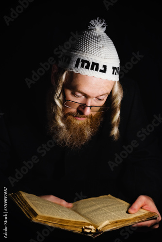 Hasidic jew reading Torah. Religious orthodox jew with sidelocks and red beard in white bale praying in the dark. Low key photo. Vertical orientation.