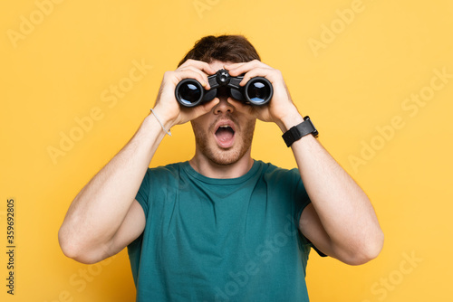 young shocked man looking through binoculars on yellow photo