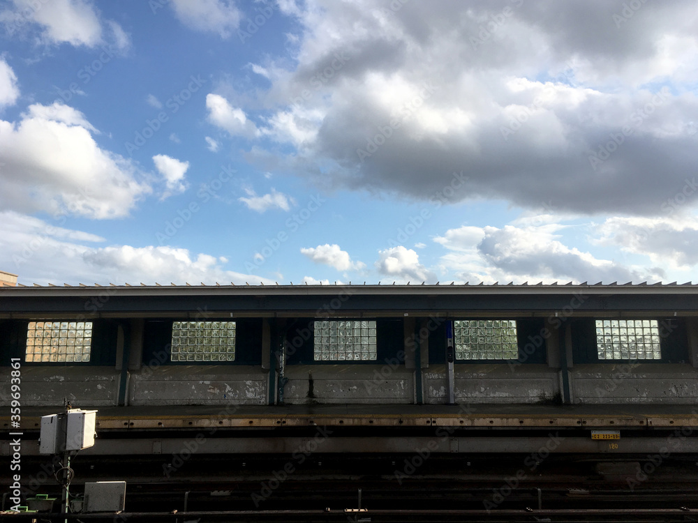 Empty overground subway platform with subway tracks in foreground. Blue sky and puffy clouds at vacant New York City train station, empty city, decline in public transport due to COVID-19.