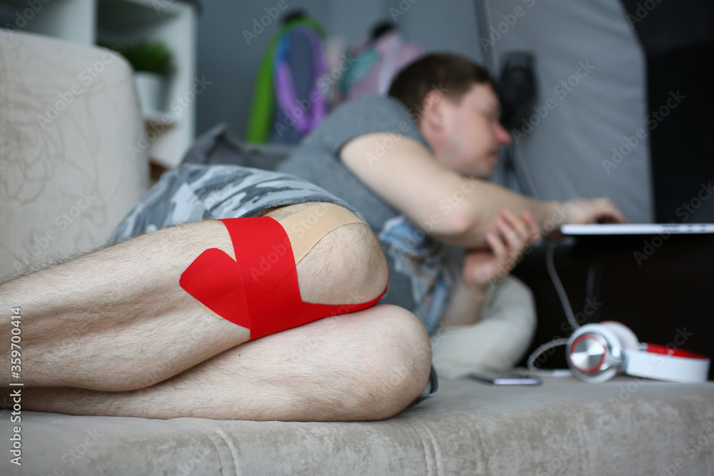 Close up of male patient with red and beige band aid on knee resting on sofa at home