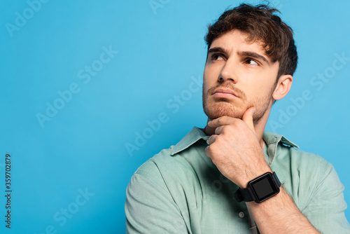 thoughtful young man looking away while touching chin on blue