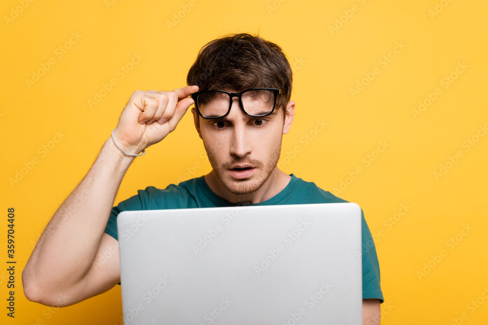 shocked young man touching glasses while using laptop on yellow shocked young man touching glasses while using laptop on yellow
