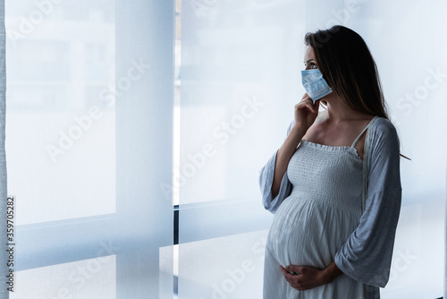 Portrait of a pregnant woman standing by the window with medical face mask, holding her belly. Worries about childbirth during pandemic.