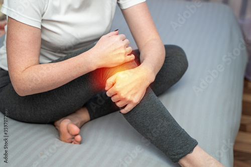 Attractive woman with pain in knee sitting on bed in apartment. Tendon knee joint problems on woman leg indicated with red spot.