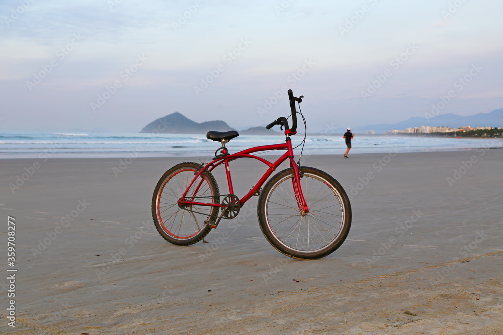 Obraz premium A red bike on the beach sand. In the background a man running. The sea, blue ocean and the horizon. Brazil, Bertioga, Sao Lourenco beach.