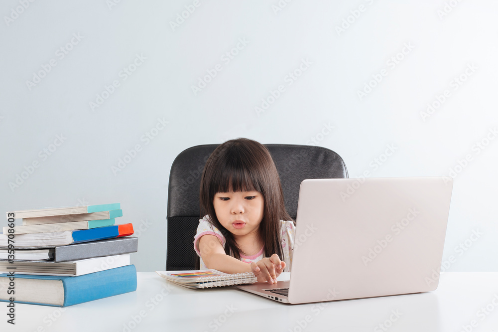 Smart asian little girl studying and doing homework using laptop on table with pile of books. Education at home concept. Isolated over grey background. Smart asian little girl studying and doing homework using laptop on table with pile of books. Education at home concept. Isolated over grey background.