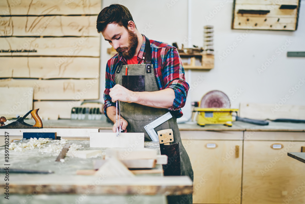 Young bearded craft master 30 years old doing wood construction during leisure time for hobby indoors, Caucasian man artisan in apron concentrated on improvement equipment furniture in workshop Young bearded craft master 30 years old doing wood construction during leisure time for hobby indoors, Caucasian man artisan in apron concentrated on improvement equipment furniture in workshop