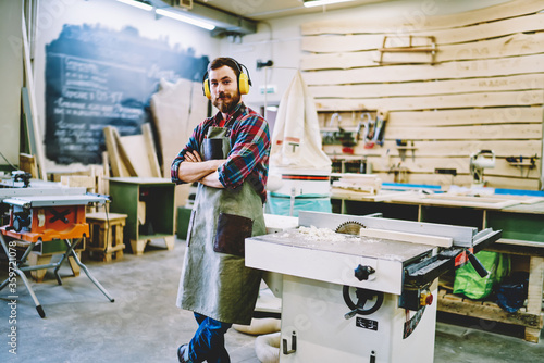 Half length portrait of self-employed woodman in work apron standing near lathe instument with crossed hands and looking at camera, successful male woodworker in protection headphones posing indoors
