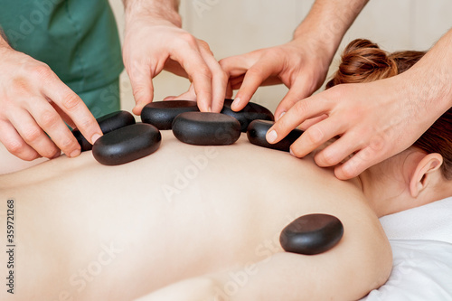 Male four hands laying large new oval black hot stones for back massage on female back in spa salon