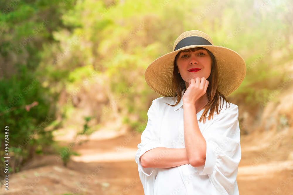 Fototapeta premium Portrait of a redhead woman in white dress and hat in the forest on a sunny day.