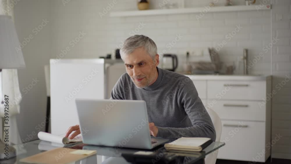 Medium shot of senior man with grey hair comparing figures in notepad and on laptop computer, closing pc and thinking with smile on his face sitting at table at home Medium shot of senior man with grey hair comparing figures in notepad and on laptop computer, closing pc and thinking with smile on his face sitting at table at home