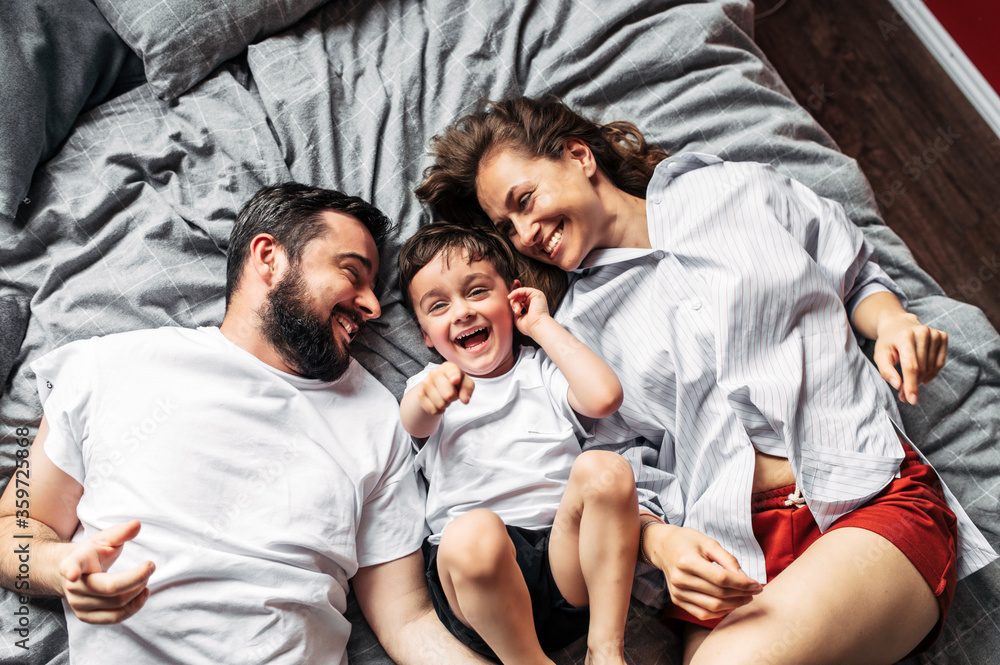 Obraz premium Happy family spend time at home together. Young parents mom and dad and their toddler son are lying on the bed in the bedroom and laughing